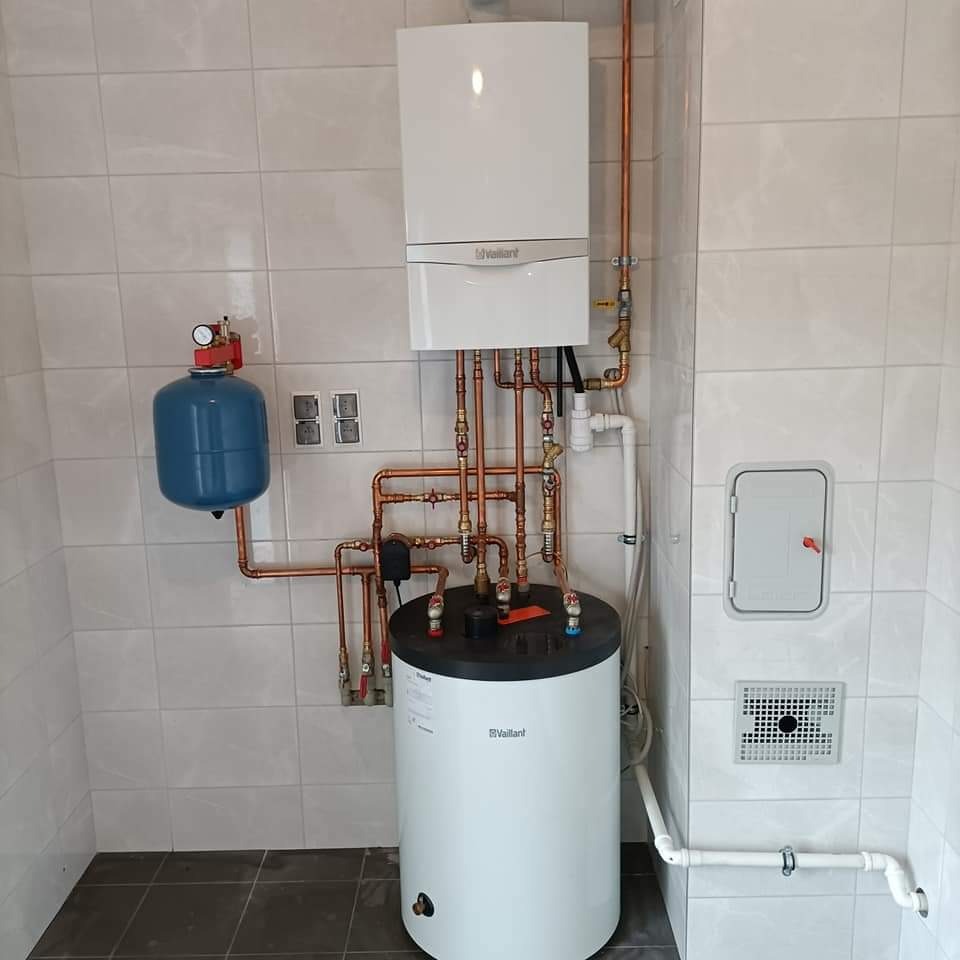 Biały piec gazowy Vaillant z podłączonym zasobnikiem i niebieskim naczyniem przeponowym na tle jasnych płytek. Miedziane rury tworzą skomplikowaną instalację.