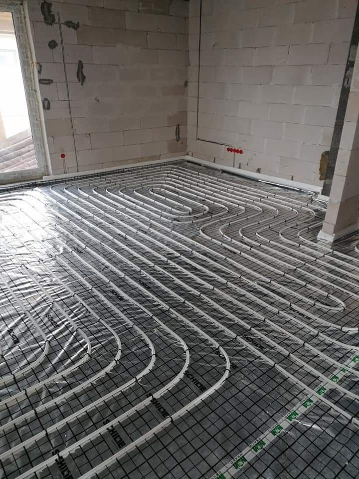 Instalacja ogrzewania podłogowego w surowym wnętrzu. Białe rury ułożone na czarnej siatce, widoczne ściany z betonu komórkowego i okno. Etap przed wylewką.