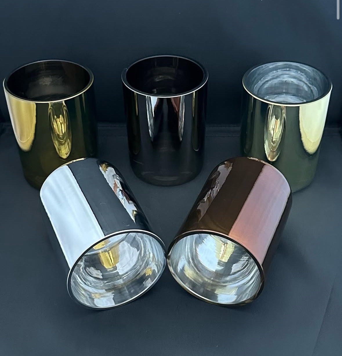 Metalowe cylindryczne formy w odcieniach złota, srebra, brązu i czerni, ustawione na ciemnym tle. Błyszczące powierzchnie odbijają światło.