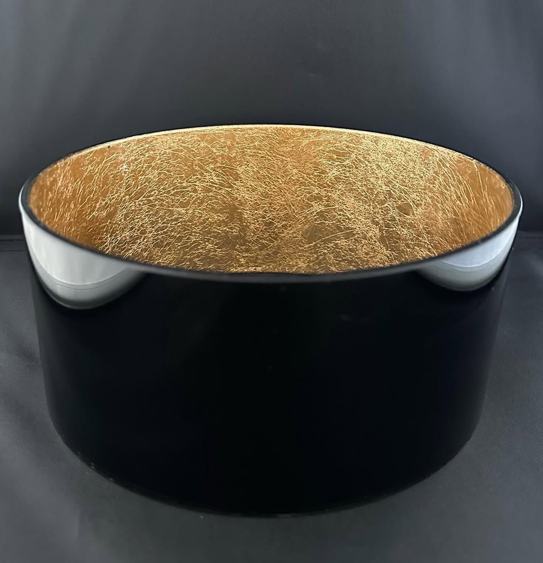 Czarna, okrągła misa z błyszczącym, złotym wnętrzem, prezentowana na ciemnym tle. Widoczny detal faktury złotej powierzchni i odbicia światła na gładkiej, czarnej obudowie.