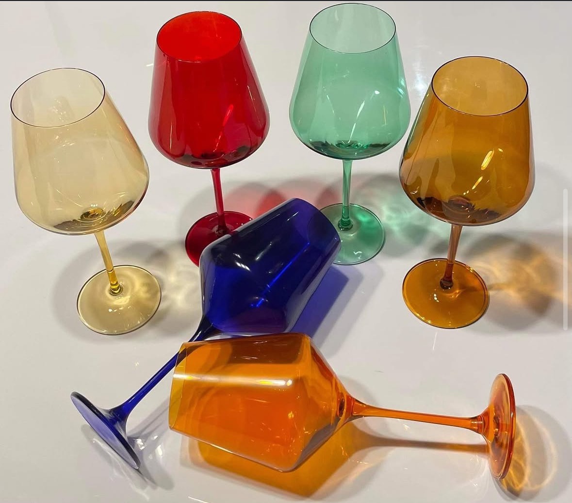Kolorowe kieliszki z barwionego szkła: żółty, czerwony, zielony, pomarańczowy i niebieski, stojące i leżące na białym tle, rzucające cienie.