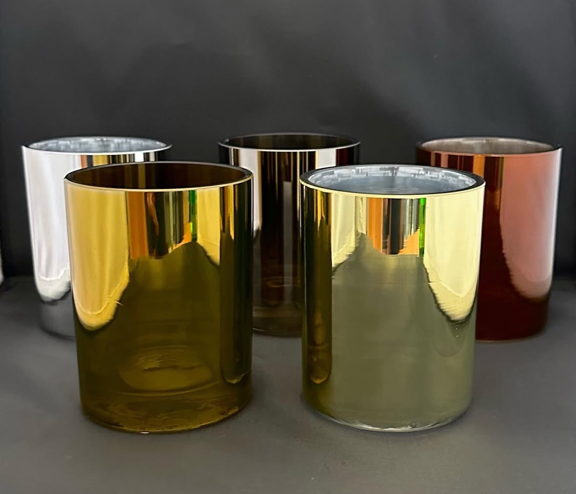 Metalizowane cylindryczne formy w odcieniach złota, srebra i miedzi, ustawione na szarym tle. Widoczne refleksy świetlne podkreślają gładką powierzchnię.