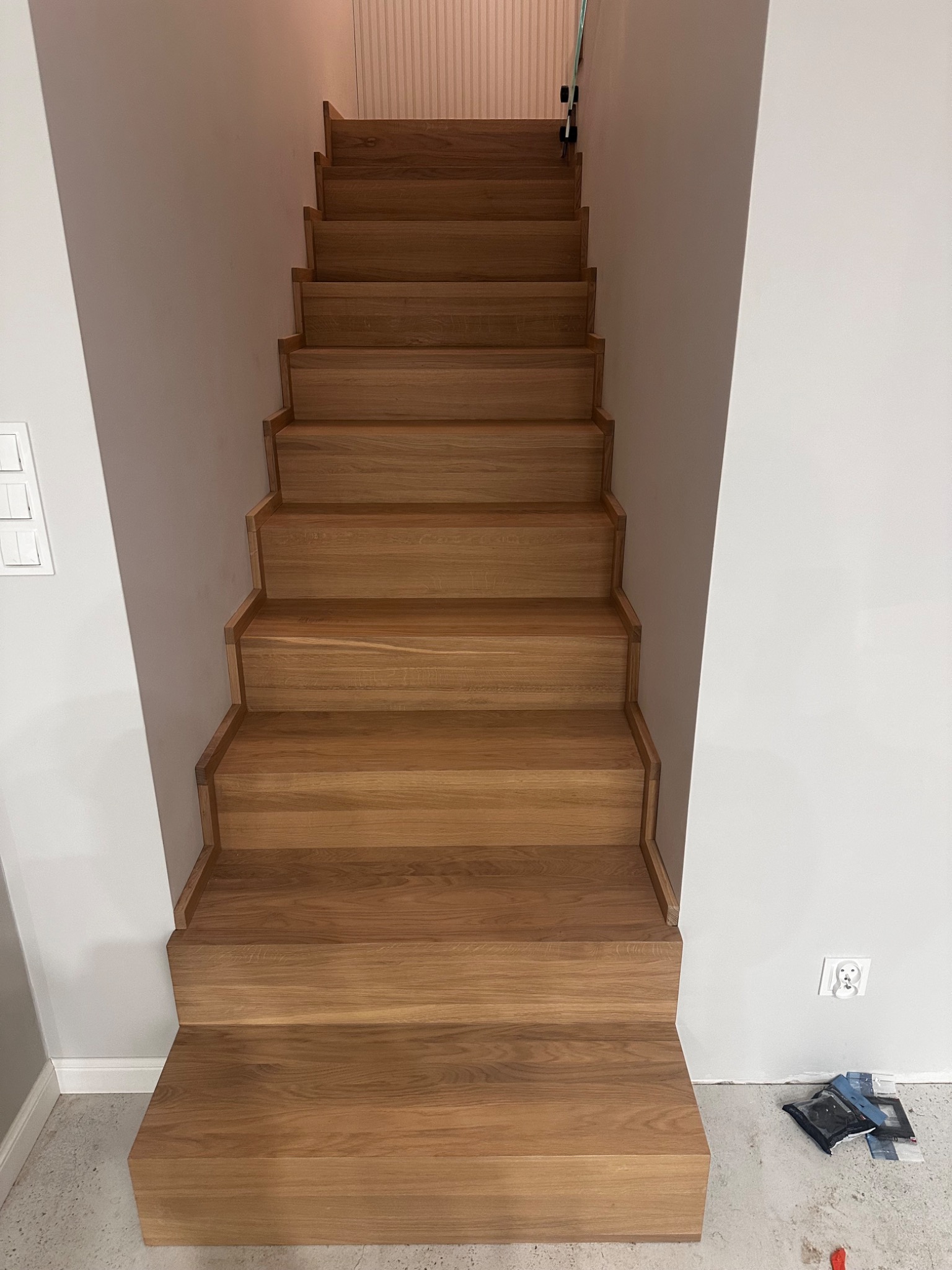 Proste, drewniane schody bez balustrady, prowadzące w górę. Widoczne stopnie z jasnego drewna, szare ściany i gniazdko elektryczne. Minimalistyczny design.