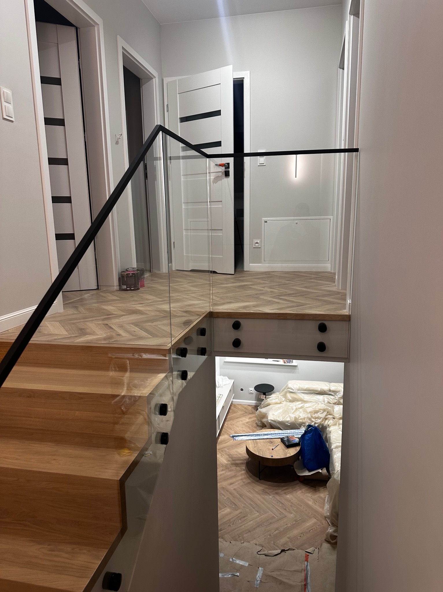 Nowoczesne schody drewniane z balustradą szklaną i czarnymi elementami mocującymi. Parkiet ułożony w jodełkę. Widok na salon w trakcie remontu.