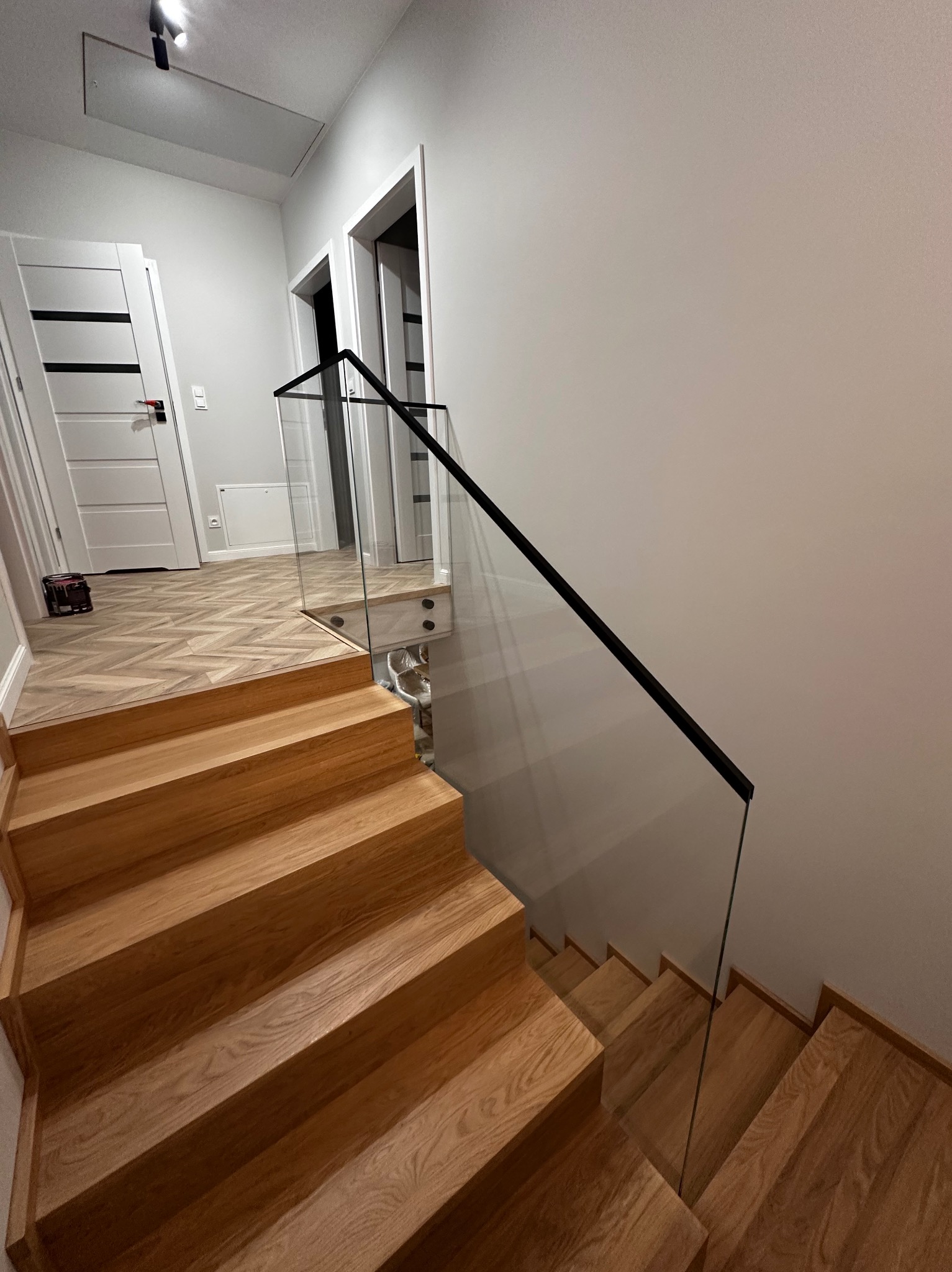 Jasne, dębowe schody z nowoczesną balustradą ze szkła i czarnych profili, prowadzące na piętro. Widoczny fragment parkietu w jodełkę i białe drzwi.