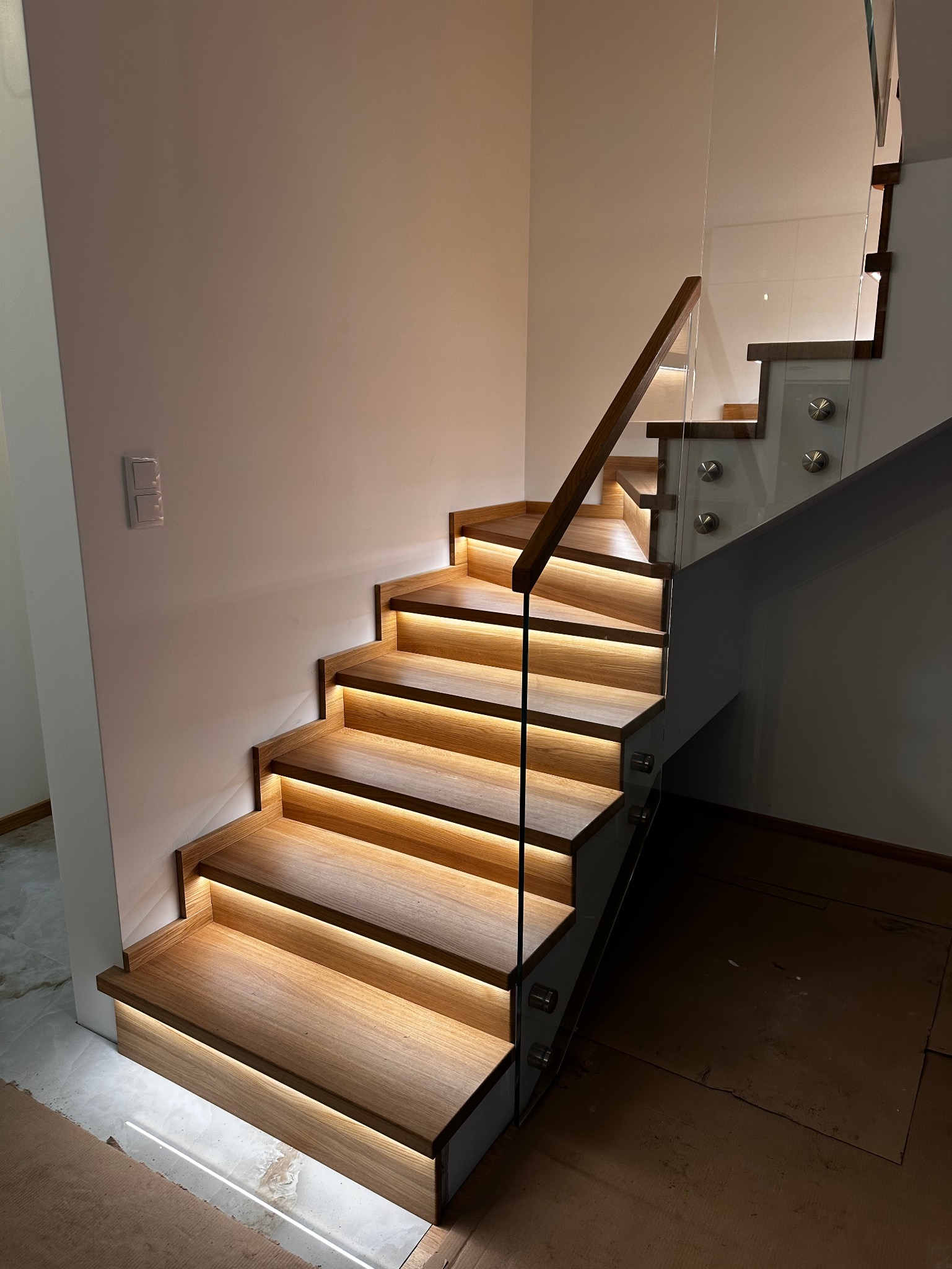 Nowoczesne schody z drewna z podświetleniem LED i szklaną balustradą. Minimalistyczny design, ciepłe światło, eleganckie wykończenie. Widok z dołu.