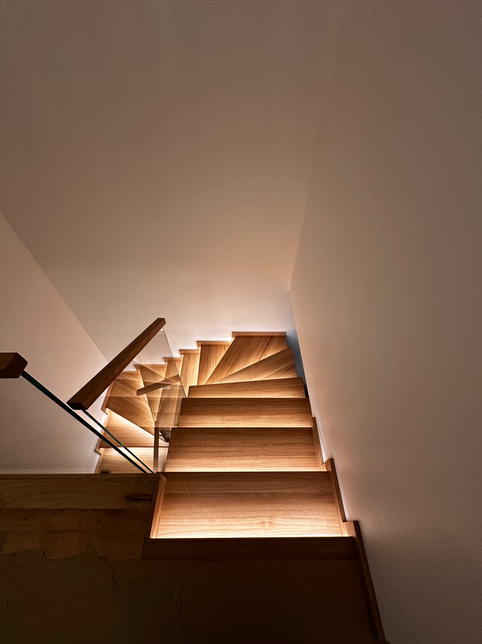 Nowoczesne schody z drewna z podświetleniem LED i szklaną balustradą, widok z góry, podkreślający spiralny kształt i ciepłe oświetlenie. Minimalistyczny design.