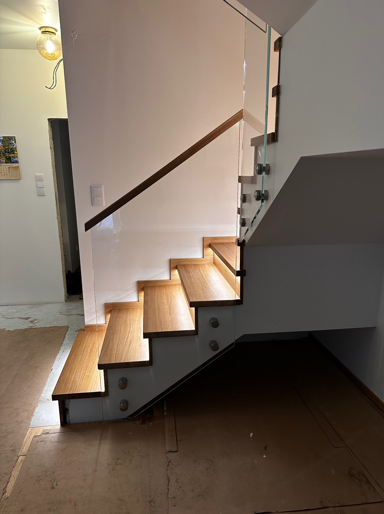 Nowoczesne schody z drewnianymi stopniami i szklaną balustradą, mocowaną punktowo. Widoczne oświetlenie LED pod stopniami i fragment pomieszczenia w tle.