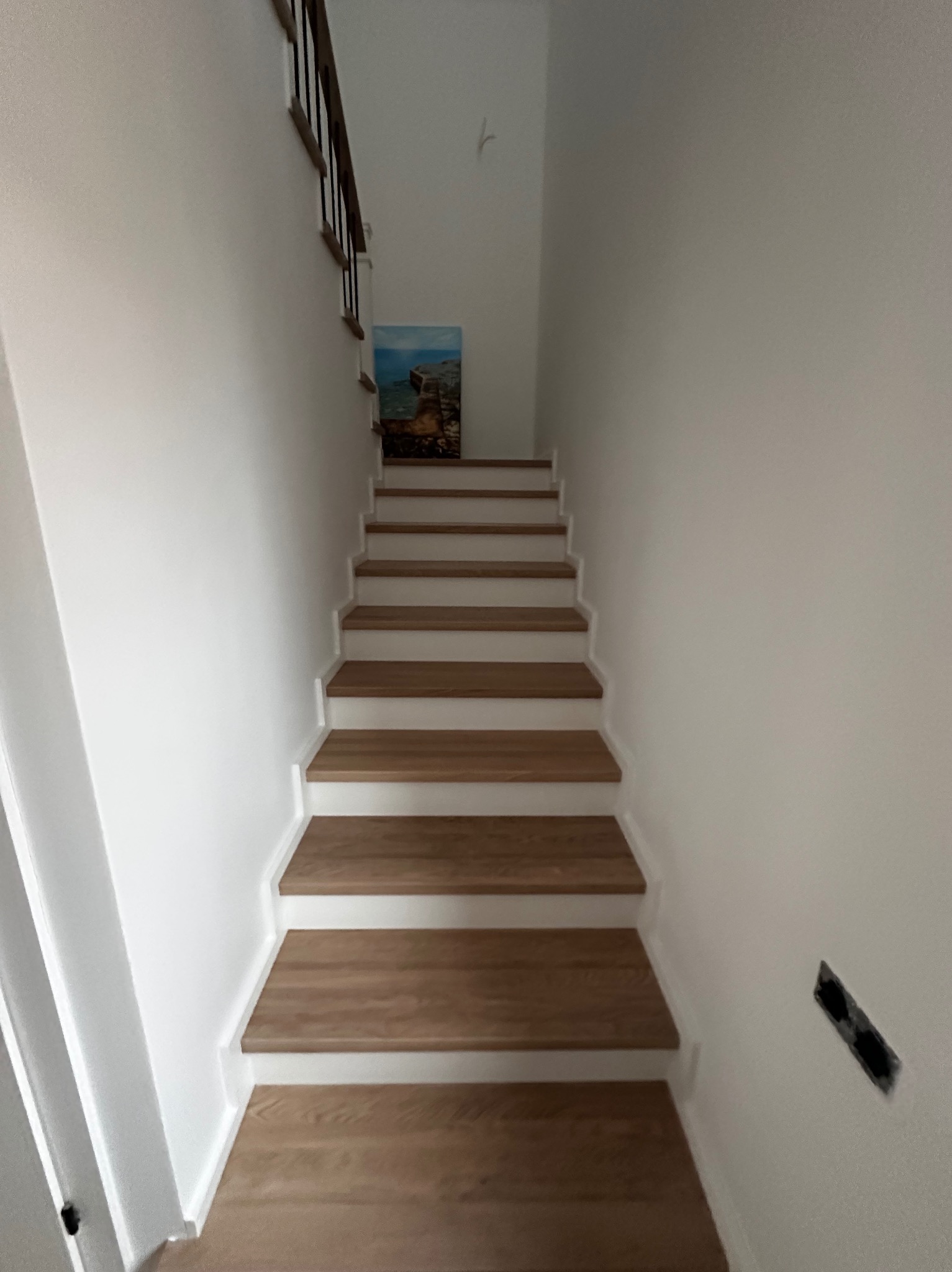 Nowoczesne schody drewniane z białymi podstopnicami i drewnianymi stopniami, widok z dołu. Na ścianie obraz z motywem morskim i niedokończone gniazdko elektryczne.