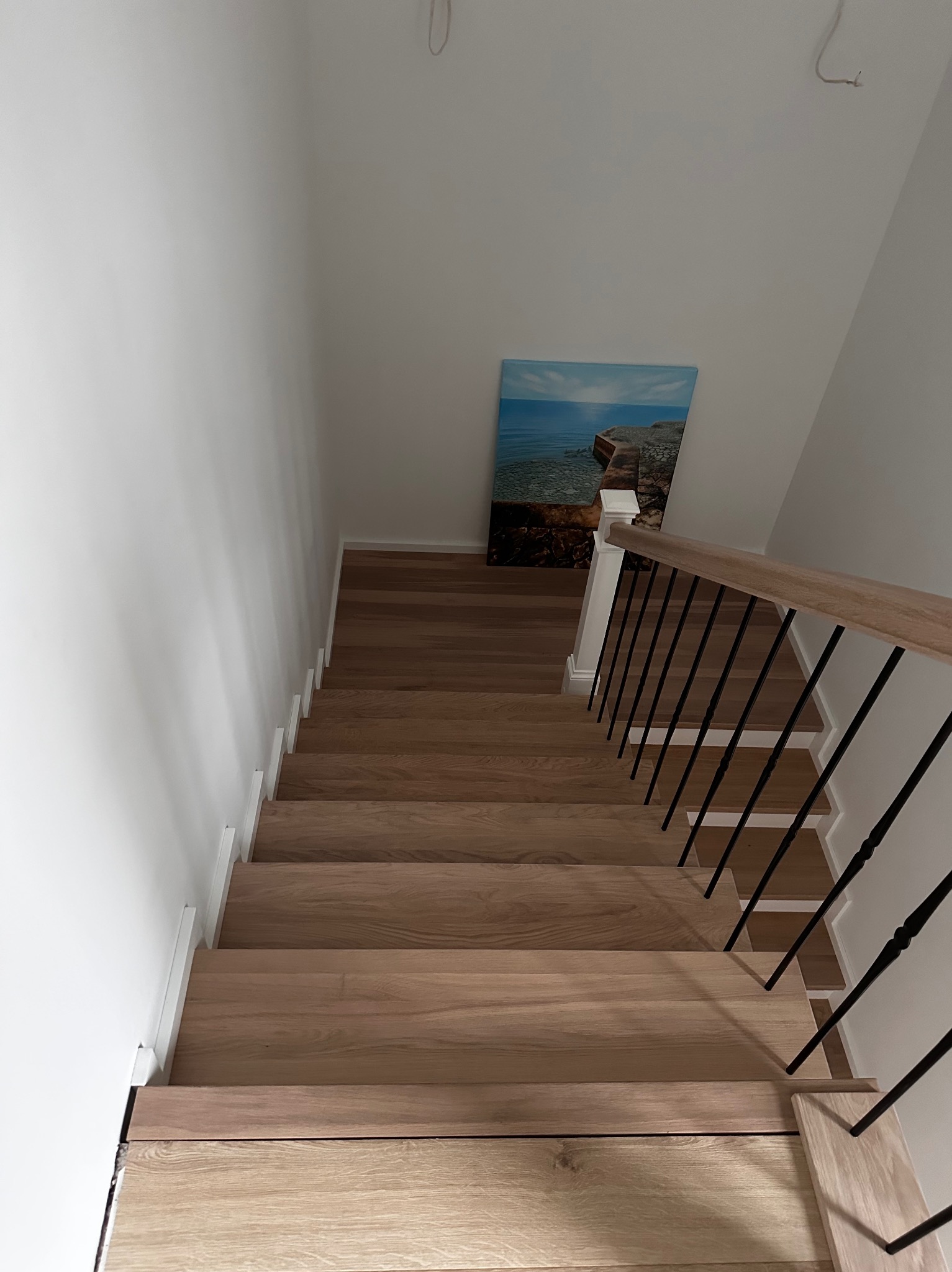 Widok z góry na jasne, drewniane schody z czarną, metalową balustradą i drewnianym poręczą. Na ścianie obraz z motywem morskim. Minimalistyczny design.