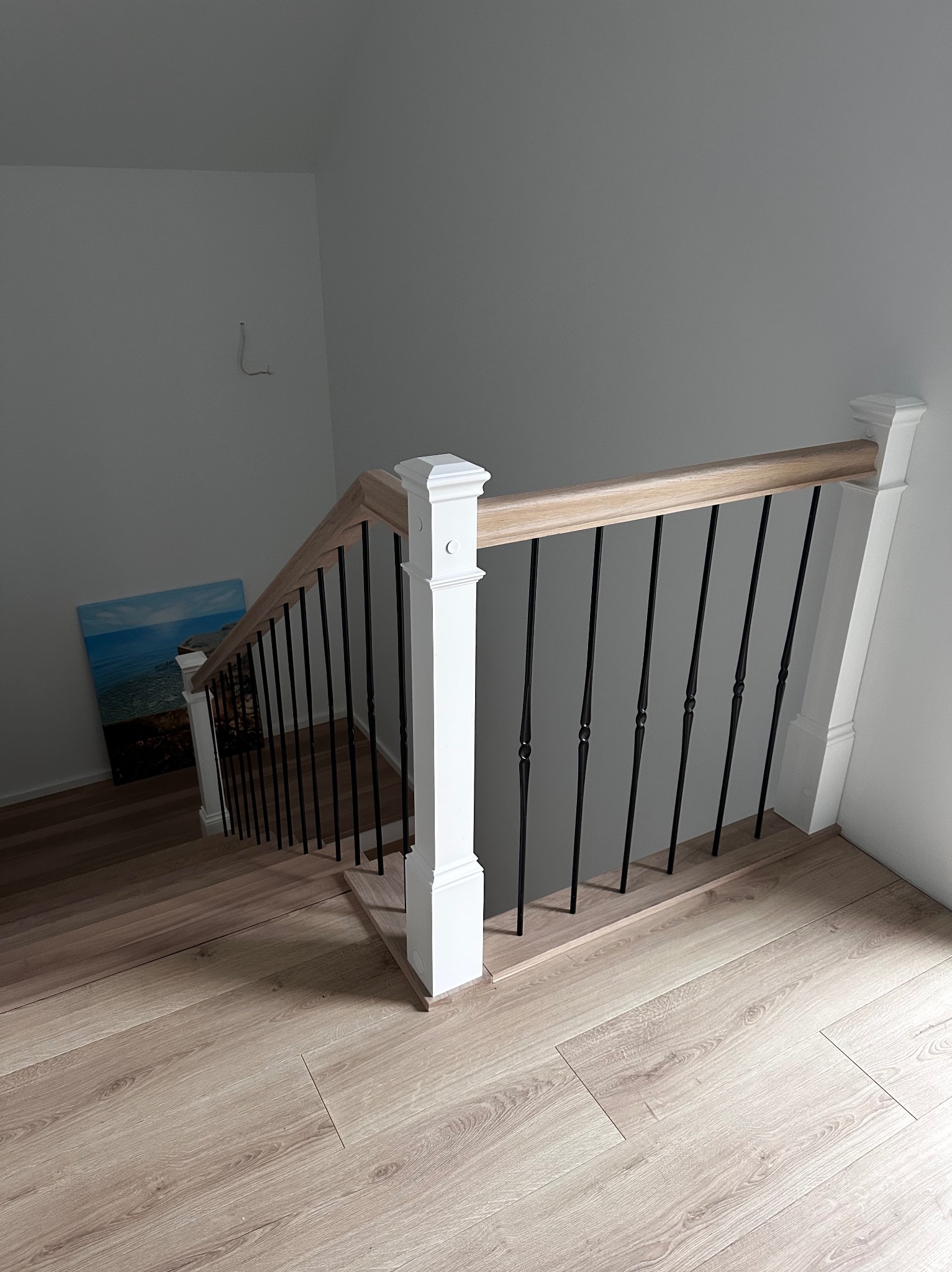 Ujęcie z góry na jasne schody z drewnianymi stopniami i białą balustradą z czarnymi, metalowymi prętami. Na ścianie w tle wisi obraz z motywem morskim.