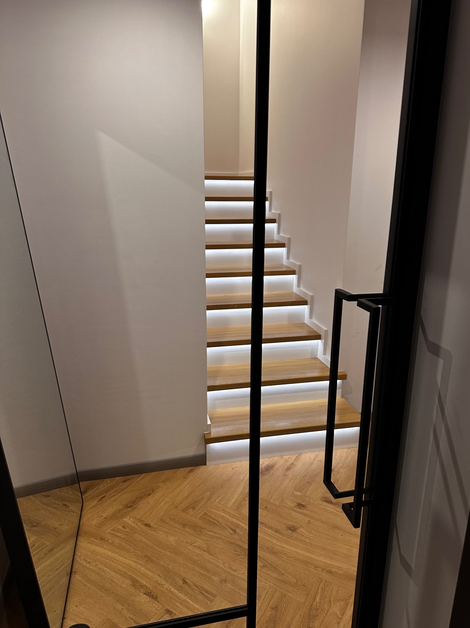 Nowoczesne schody z podświetleniem LED, widoczne przez czarną, metalową balustradę. Parkiet w jodełkę na podłodze. Minimalistyczny design.