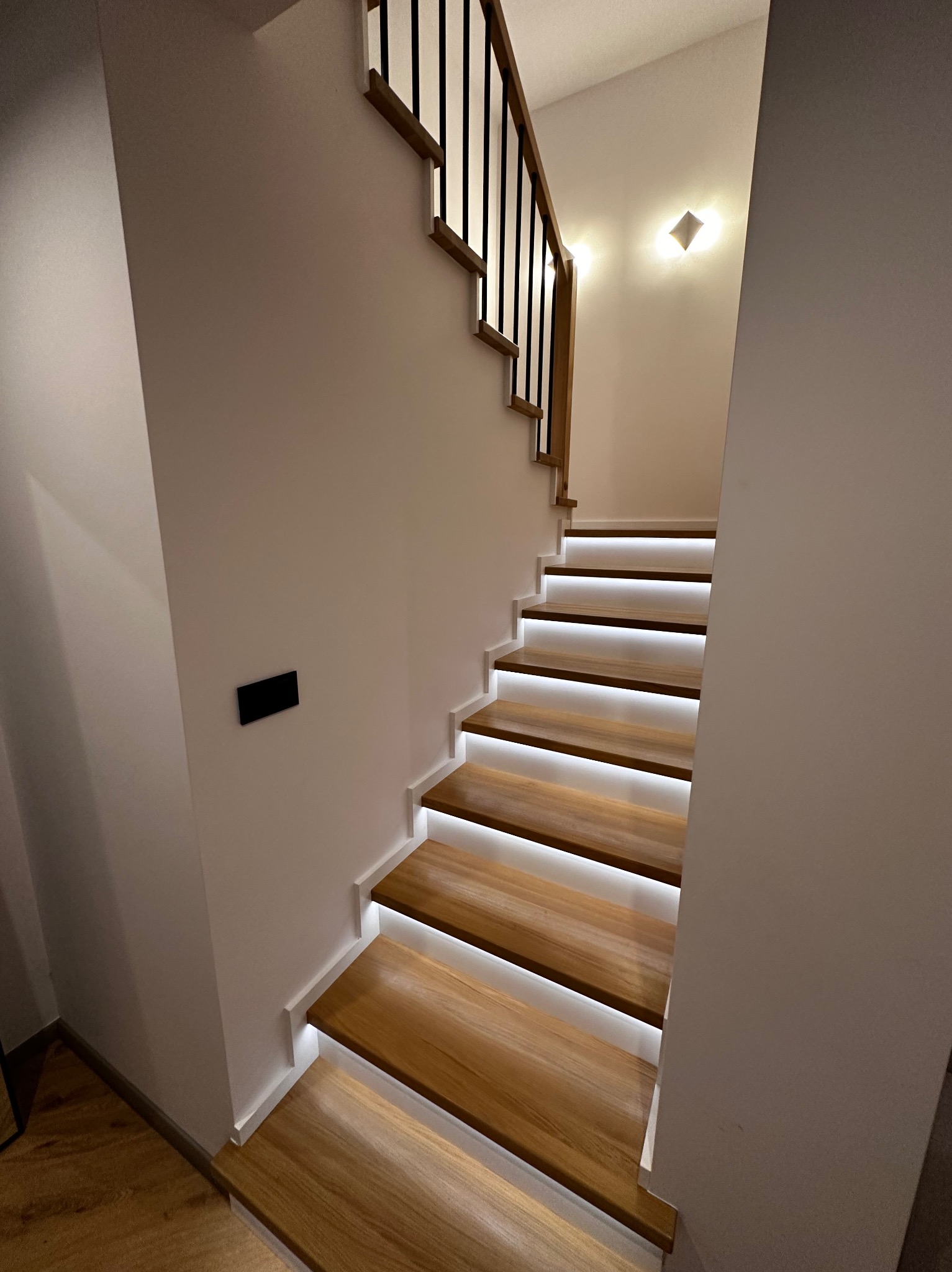 Nowoczesne schody z podświetleniem LED i czarną balustradą. Jasne drewno stopni kontrastuje z białymi ścianami. Minimalistyczny design, oświetlenie punktowe.