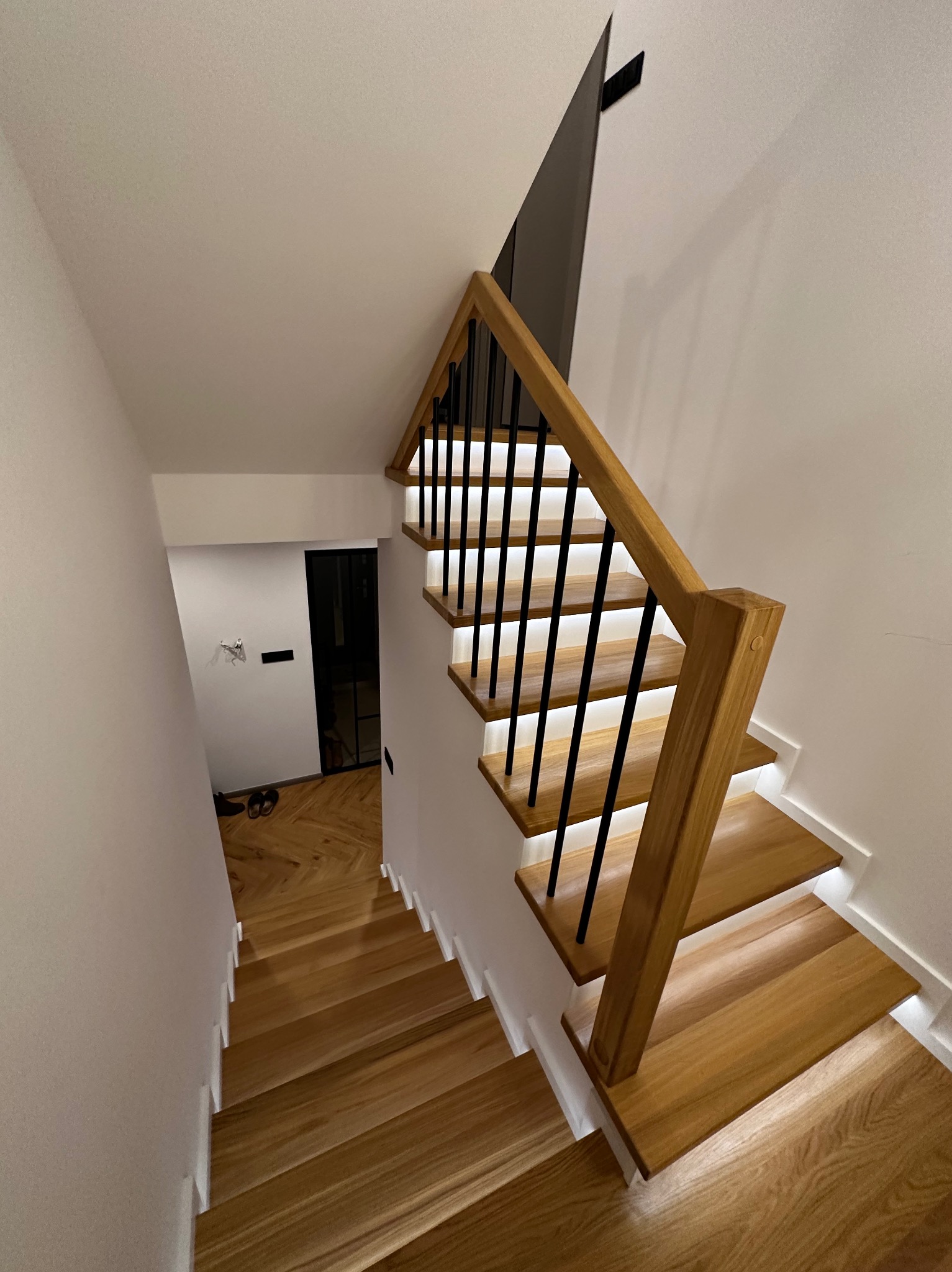 Nowoczesne schody z drewna z czarną balustradą i subtelnym oświetleniem LED, widok z góry. Parkiet w jodełkę widoczny na dole schodów.