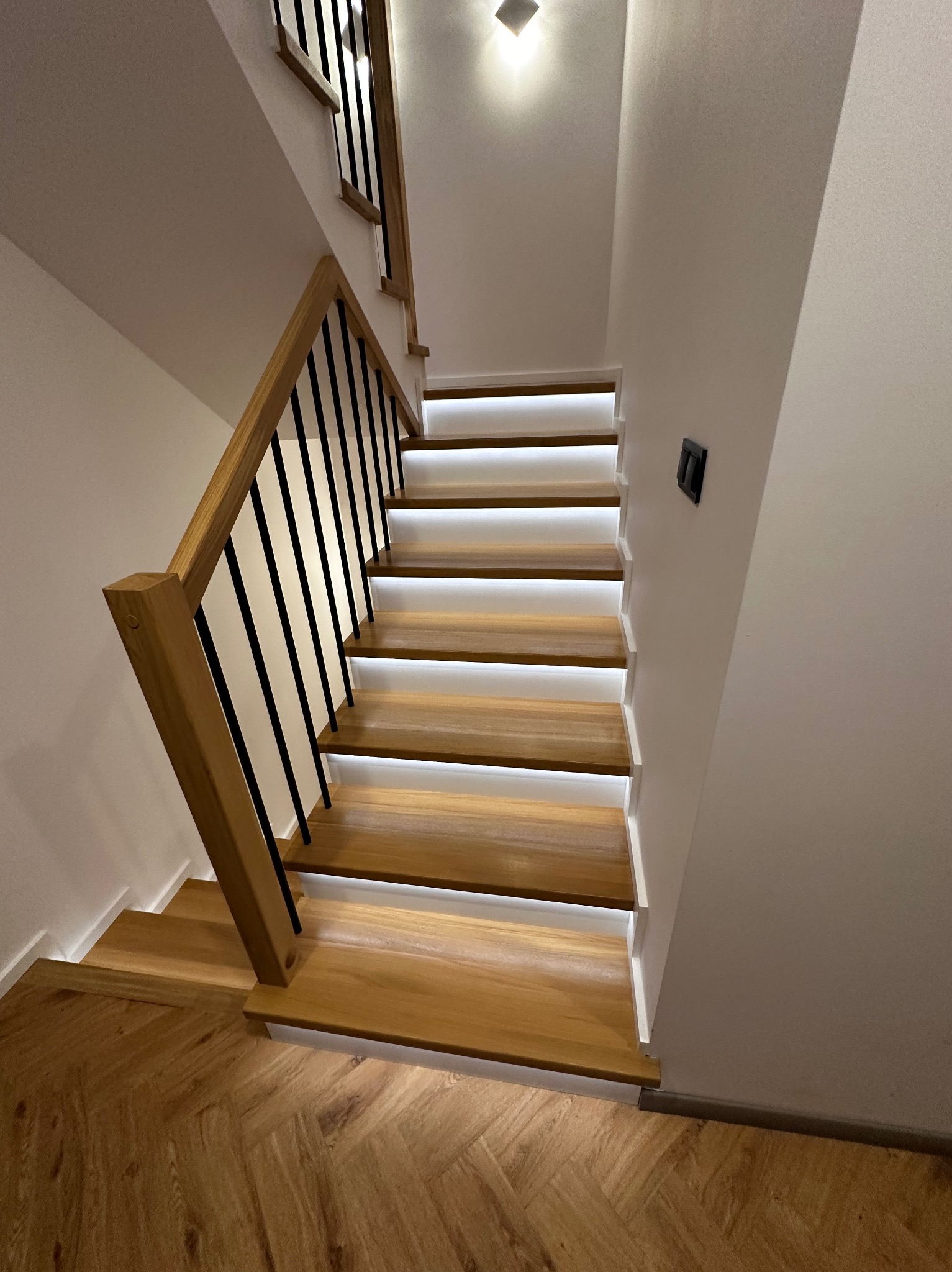 Nowoczesne schody z drewna z podświetleniem LED, czarne metalowe balustrady, parkiet w jodełkę. Ujęcie z dołu, widok na stopnie i fragment ściany z włącznikiem.