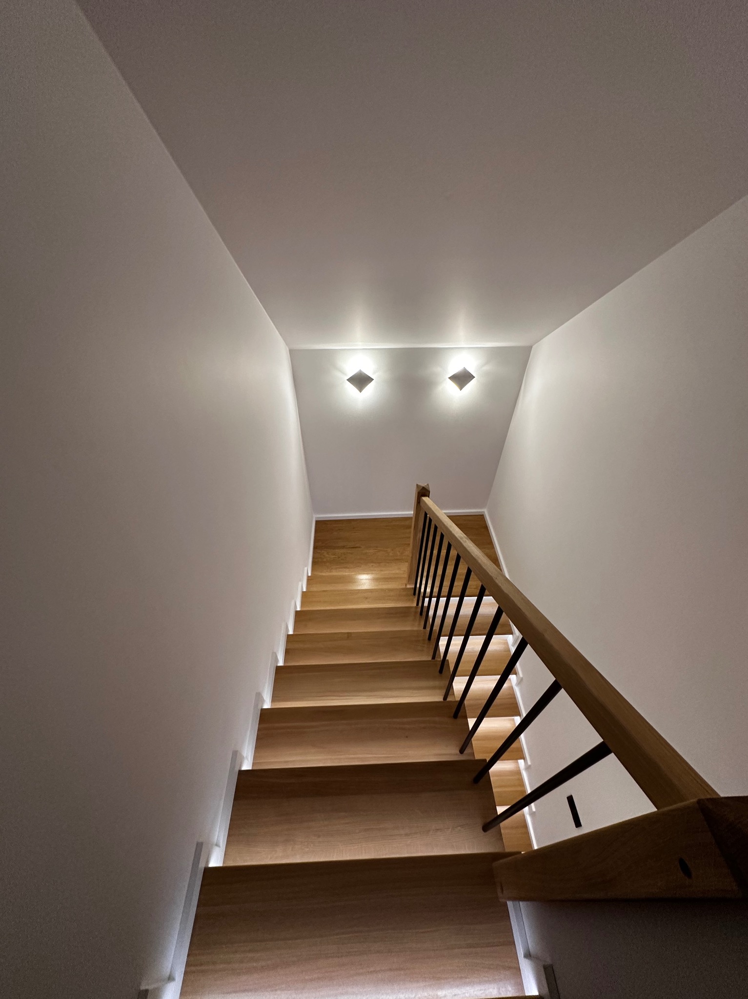 Nowoczesne schody drewniane z czarną balustradą i subtelnym oświetleniem LED wzdłuż stopni, widziane z dołu, podkreślające minimalistyczny design.