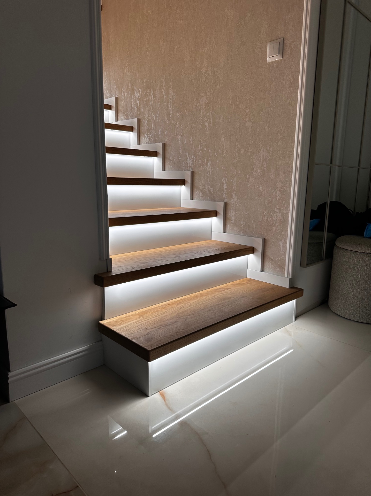 Nowoczesne schody z drewnianymi stopniami i białymi podstopnicami, podświetlone ledowym światłem, na tle jasnej ściany i błyszczącej podłogi.