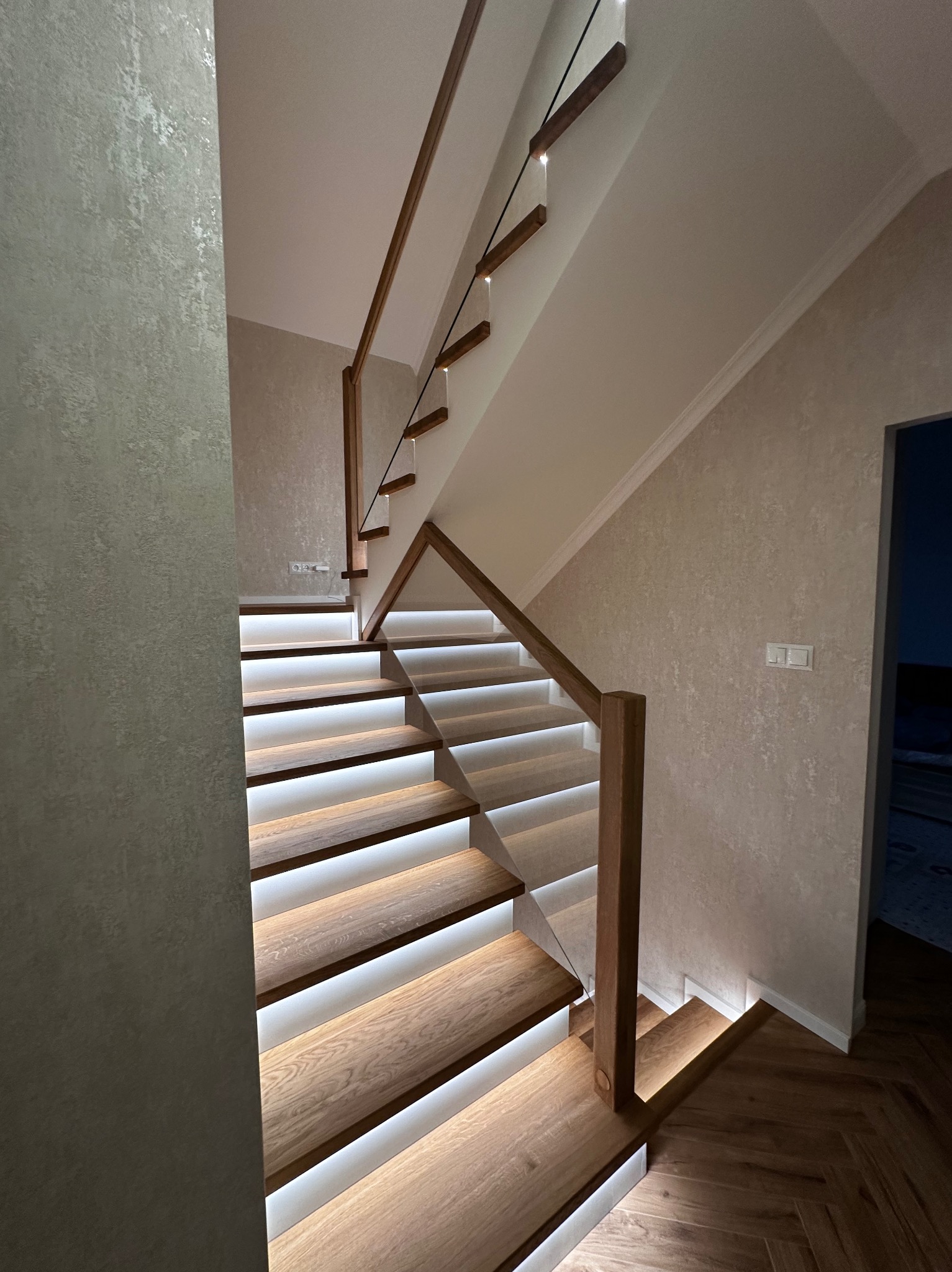 Nowoczesne schody z podświetleniem LED, drewniane stopnie i szklana balustrada z drewnianymi słupkami. Parkiet w jodełkę na podłodze, ściany w jasnym odcieniu.