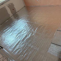 K.E.M remonty i wykończenia - Błyszcząca folia grzewcza rozłożona na podłodze w trakcie remontu poddasza. Widoczne okno dachowe, fragment ściany ze śladami szpachlowania i przewody elektryczne.