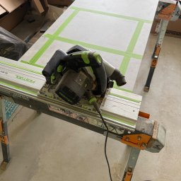 K.E.M remonty i wykończenia - Cięcie płyty pilarką Festool na stole roboczym z zieloną taśmą wyznaczającą linie cięcia. Widoczne kartony w tle. Ujęcie z góry, bliski plan na narzędzie.