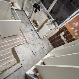 K.E.M remonty i wykończenia - Układanie szarych płytek na podłodze z ogrzewaniem podłogowym, widoczne krzyżyki dystansowe i częściowo zamontowane białe szafki kuchenne. Ukończony etap prac.