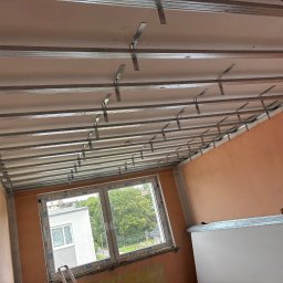 K.E.M remonty i wykończenia - Stelaż sufitu podwieszanego z profili metalowych, widok z dołu na tle okna i pomarańczowych ścian. Elementy montażowe widoczne. Etap prac wykończeniowych.