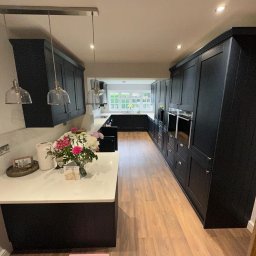 K.E.M remonty i wykończenia - Elegancka, czarna zabudowa kuchenna z białym blatem i kwiatami. Widok na dalszą część kuchni z oknem. Nowoczesne lampy wiszące nad blatem.