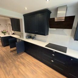 K.E.M remonty i wykończenia - Nowoczesna kuchnia z ciemnymi szafkami, białym blatem i drewnianą podłogą. Otwarta szafka ukazuje wnętrze. Widoczny okap i płyta indukcyjna.