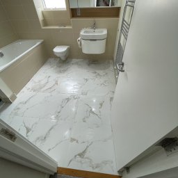 K.E.M remonty i wykończenia - Jasna łazienka z marmurową podłogą, widok zza uchylonych drzwi. Umywalka, toaleta i grzejnik drabinkowy. W lustrze odbija się osoba robiąca zdjęcie.