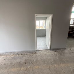 K.E.M remonty i wykończenia - Szare ściany po malowaniu, białe drzwi otwarte do pokoju z oknem. Surowa podłoga przed wykończeniem. Widok na wnętrze po odświeżeniu.