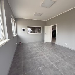 K.E.M remonty i wykończenia - Wnętrze pomalowane na szaro z szarą podłogą z płytek. Widoczne okna, drzwi i oświetlenie sufitowe. Minimalistyczny design, świeżo po remoncie.