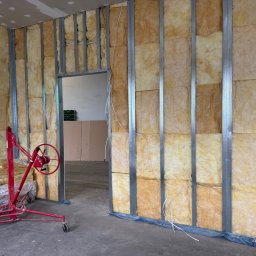 K.E.M remonty i wykończenia - Montaż izolacji z wełny mineralnej w metalowym stelażu ścianki działowej, widoczny podnośnik do płyt i fragment sufitu podwieszanego z płyt gipsowych.