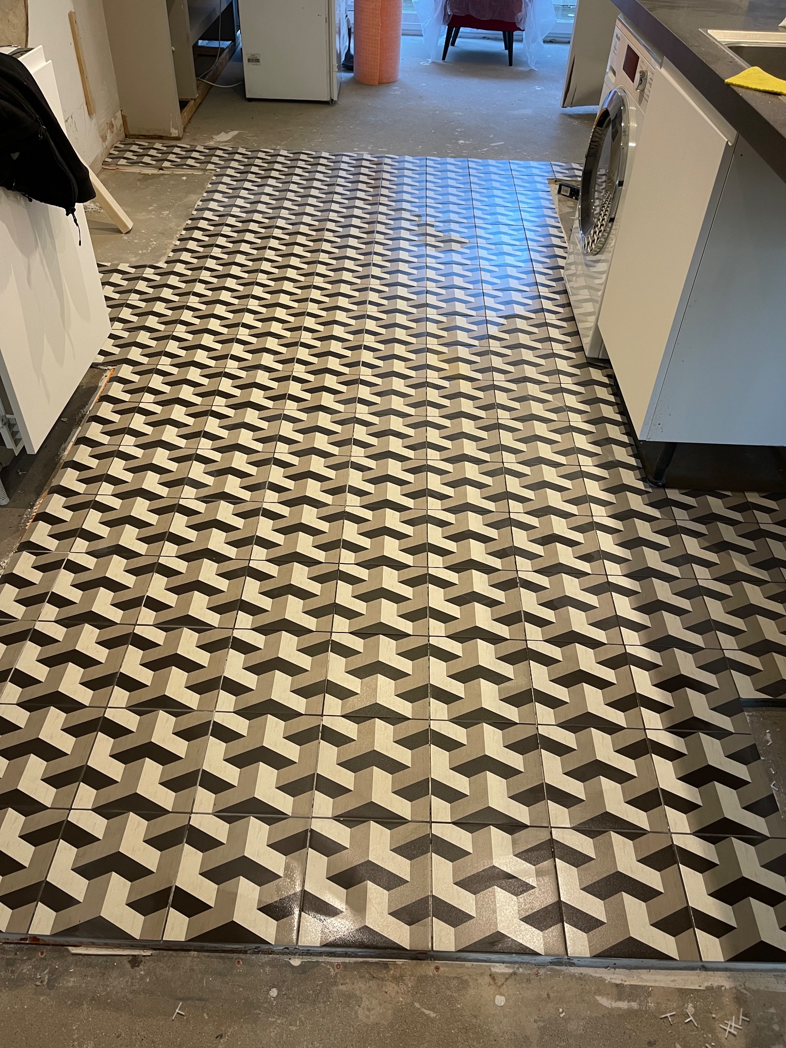 Ułożona podłoga z płytek o wzorze 3D w odcieniach szarości i beżu. Widoczna pralka i fragmenty niedokończonej podłogi. Ujęcie z góry, perspektywa podłogi.