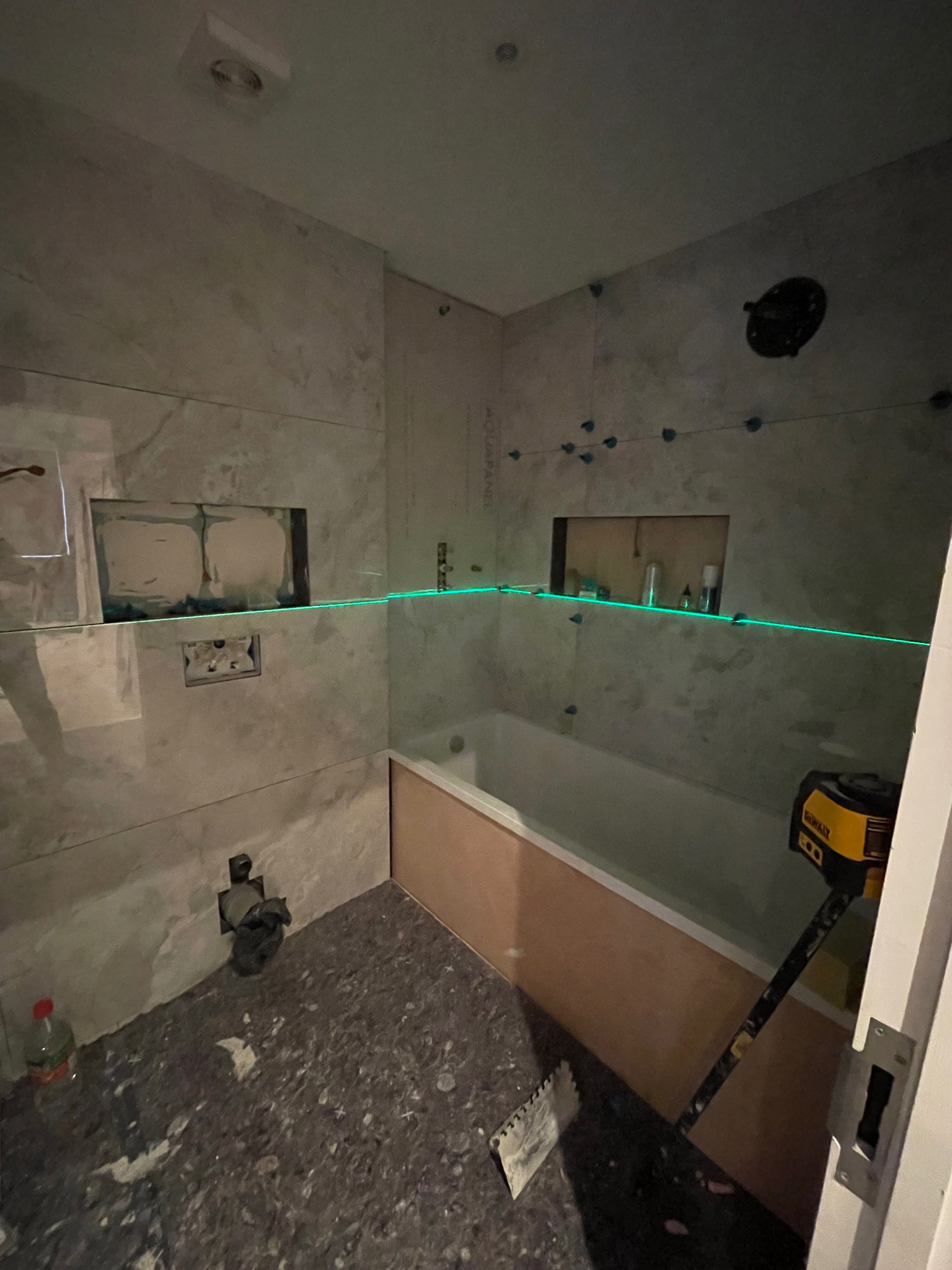 Łazienka w trakcie prac: ułożone płytki imitujące marmur, poziomica laserowa wyznacza linię, widoczne wnęki w ścianie, wanna obudowana płytą. Podłoga przygotowana pod wylewkę.