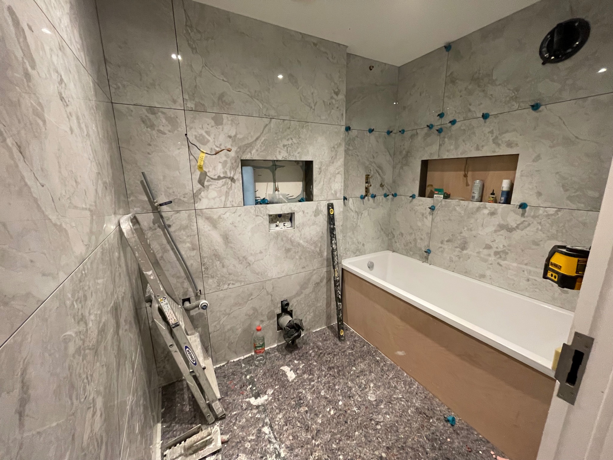 Układanie płytek w łazience w Krosno Odrzańskie. Widoczna wanna, drabina i poziomica. Płytki imitujące marmur, niebieskie krzyżyki dystansowe.