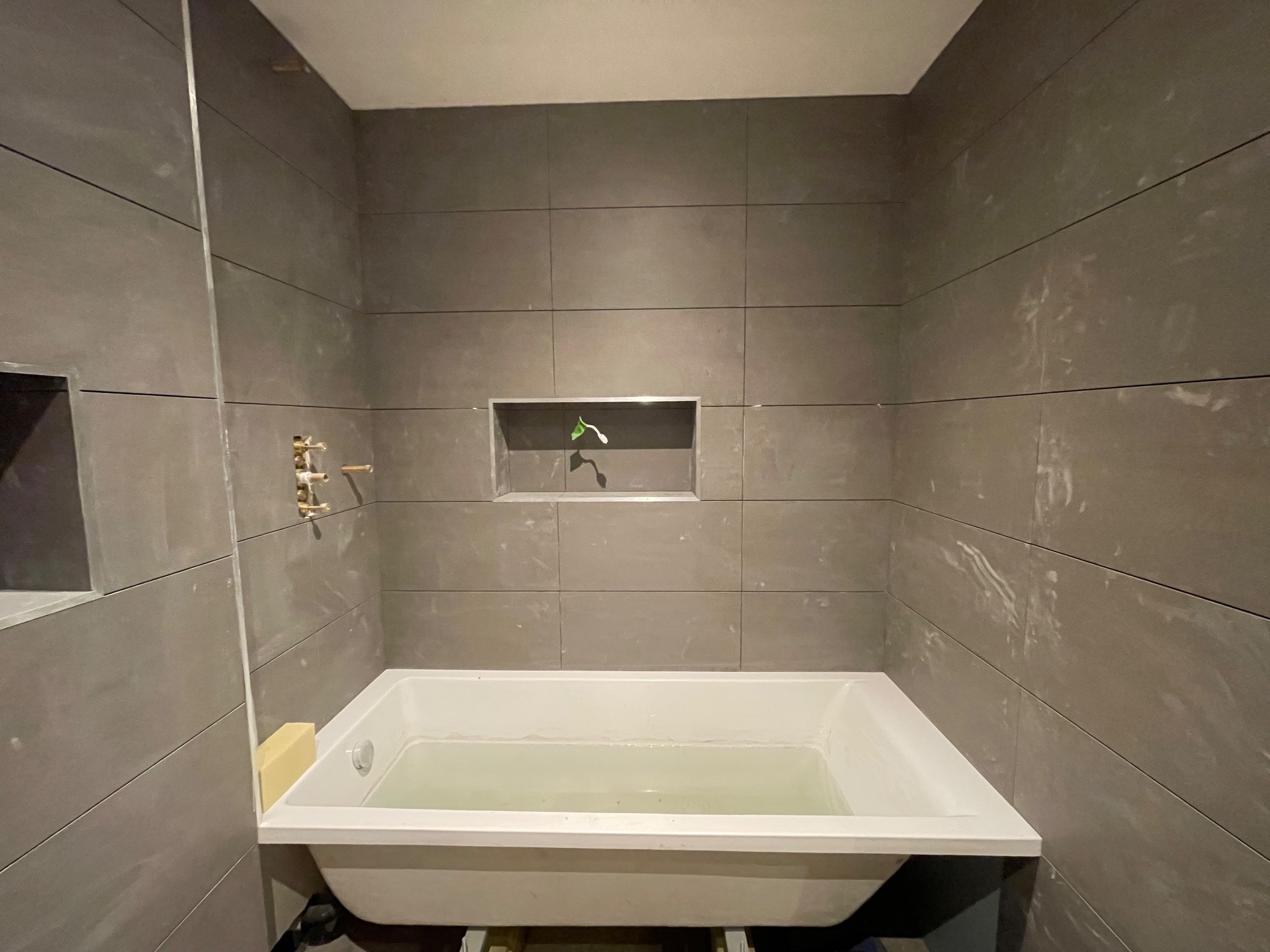 Łazienka w trakcie remontu: szare, poziome płytki, biała wanna wypełniona wodą, wnęka z zieloną rośliną. Widoczne ślady po kleju na płytkach.