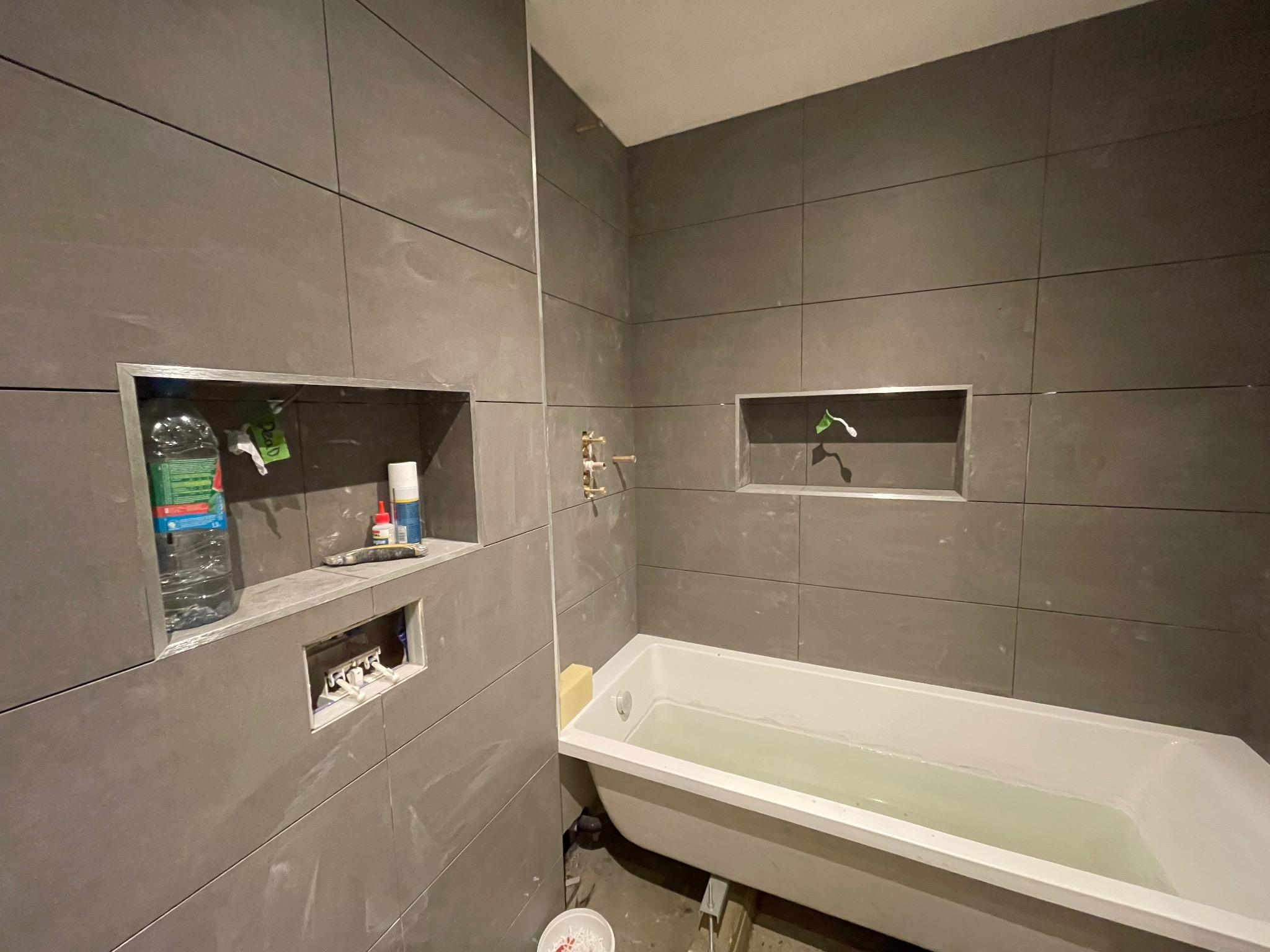 Łazienka w trakcie wykończenia: szare, prostokątne płytki, wnęki w ścianie, biała wanna wypełniona wodą. Widoczne narzędzia i materiały budowlane.