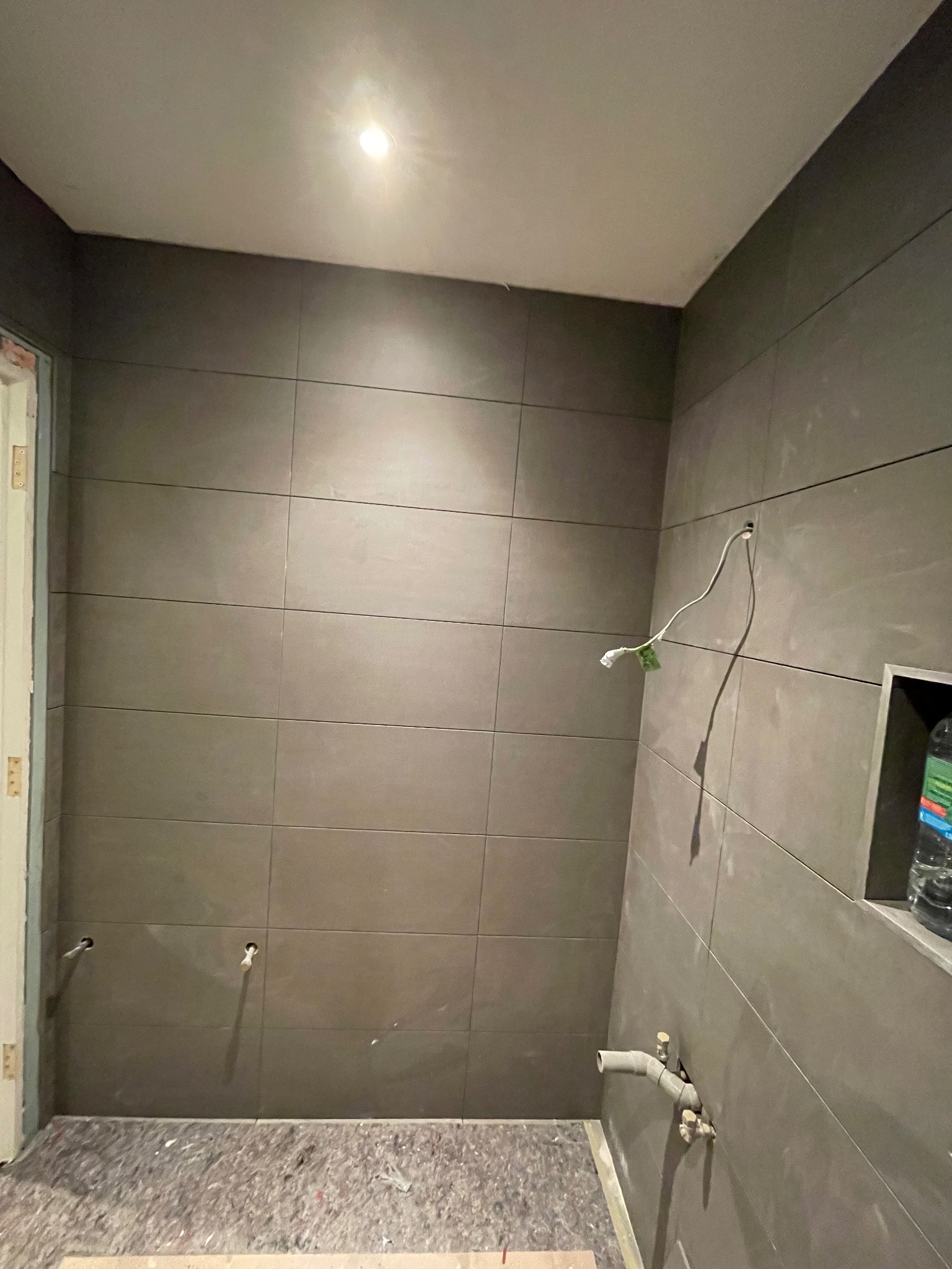 Wnętrze łazienki w trakcie prac wykończeniowych. Widoczne szare płytki na ścianach, wystające rury i przewody. Podłoga zabezpieczona materiałem budowlanym.