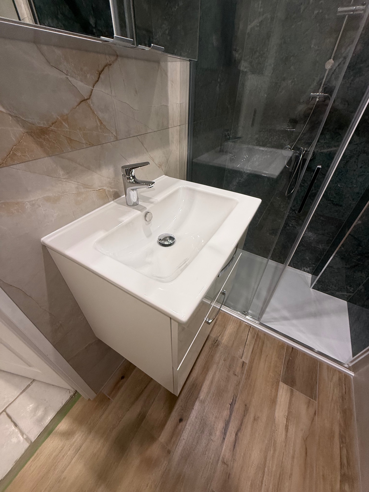 Nowoczesna łazienka po remoncie: biała umywalka z szafką, chromowana bateria, kabina prysznicowa z ciemnymi płytkami, podłoga imitująca drewno.