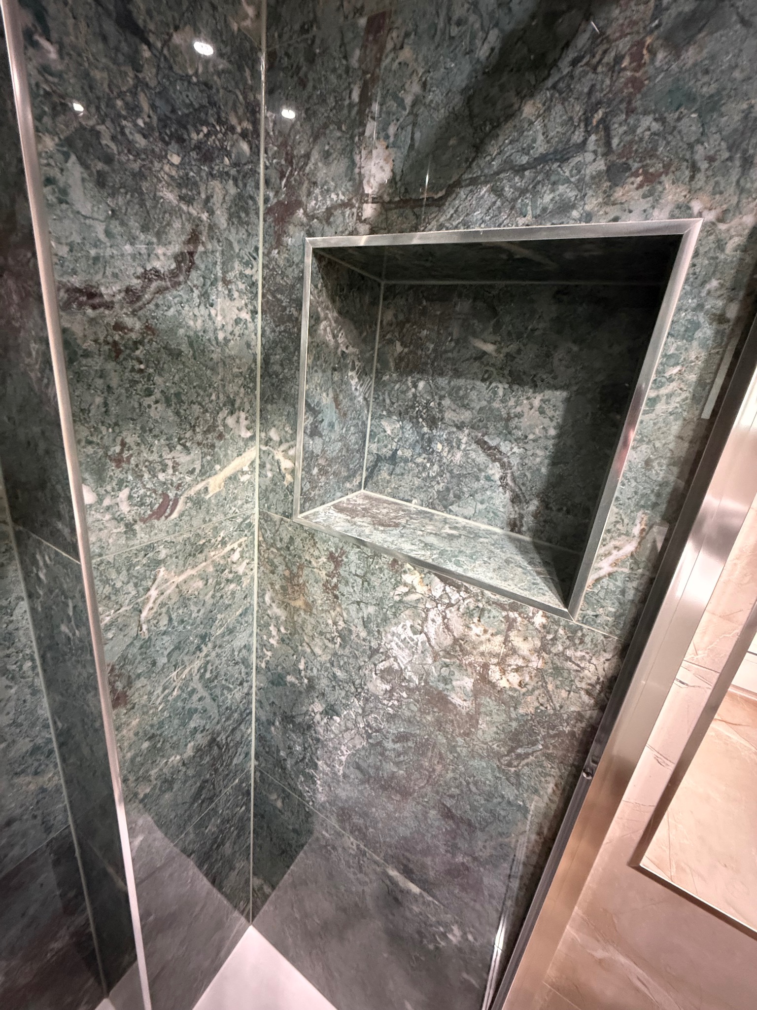 Narożna półka w kabinie prysznicowej wyłożonej zielonym marmurem z wyraźnym wzorem żyłkowania. Metalowe wykończenia podkreślają nowoczesny design.