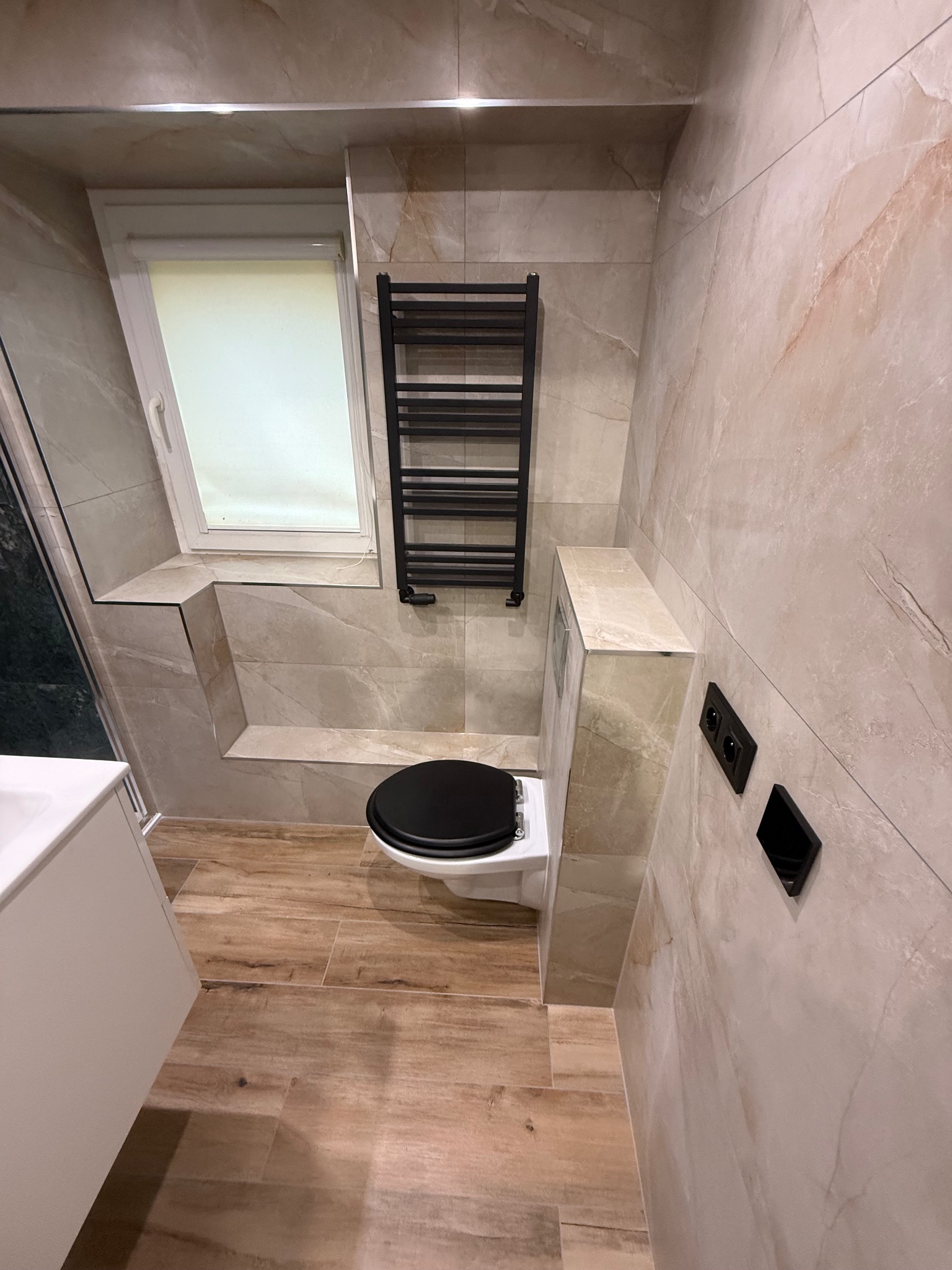 Wykończona łazienka z beżowymi płytkami imitującymi marmur, drewnopodobną podłogą, czarnym grzejnikiem drabinkowym i oknem dachowym. Nowoczesne gniazdka w czarnych ramkach.