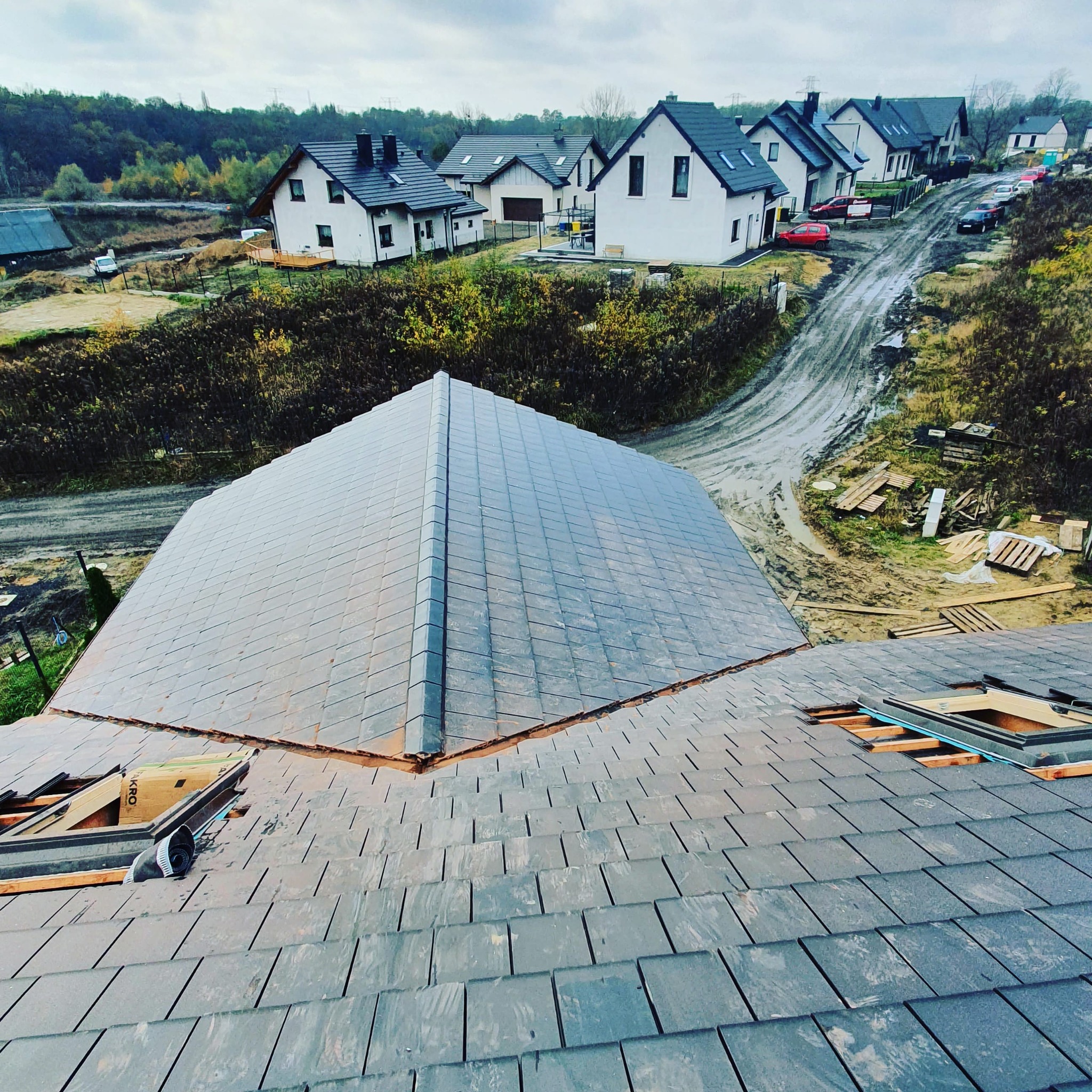 Układanie grafitowej dachówki na dachu domu jednorodzinnego. Widoczne okna dachowe w trakcie montażu i fragmenty niezakończonego krycia. W tle zabudowa osiedlowa.