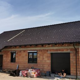 Be-Gro Dachy - Dom w trakcie budowy z ciemnobrązowym dachem z dachówki ceramicznej. Widoczne rynny i fragment drewnianej łaty na dachu. Na pierwszym planie materiały budowlane.