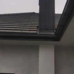 Be-Gro Dachy - Szary dach z dachówki ceramicznej i komin z blachy na tle pochmurnego nieba. Widoczna ściana budynku z oknem i rynna.