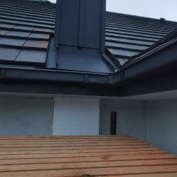 Be-Gro Dachy - Nowy, grafitowy komin z blachy na tle dachu z dachówki ceramicznej i szarego nieba. Widoczny fragment rusztowania.