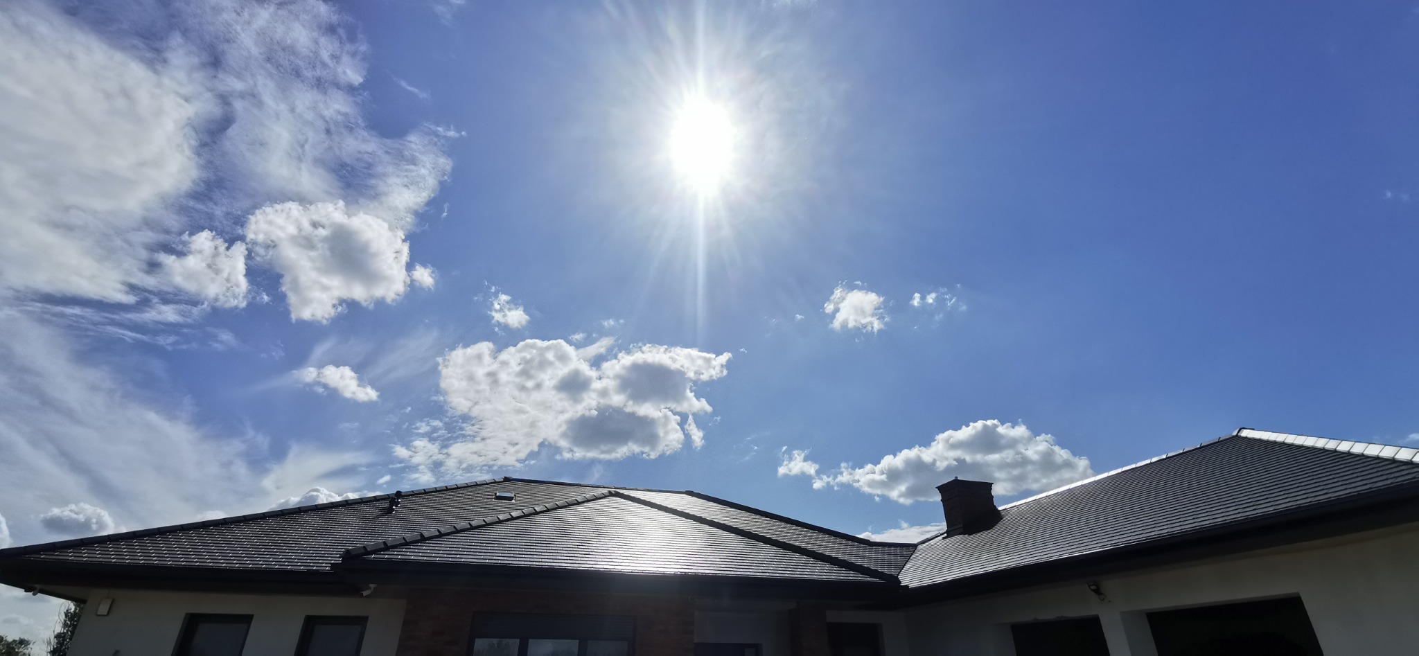 Błyszczący, grafitowy dach z dachówki ceramicznej pod błękitnym niebem z chmurami i promieniami słońca. Widok z dołu na wykończony dach domu.