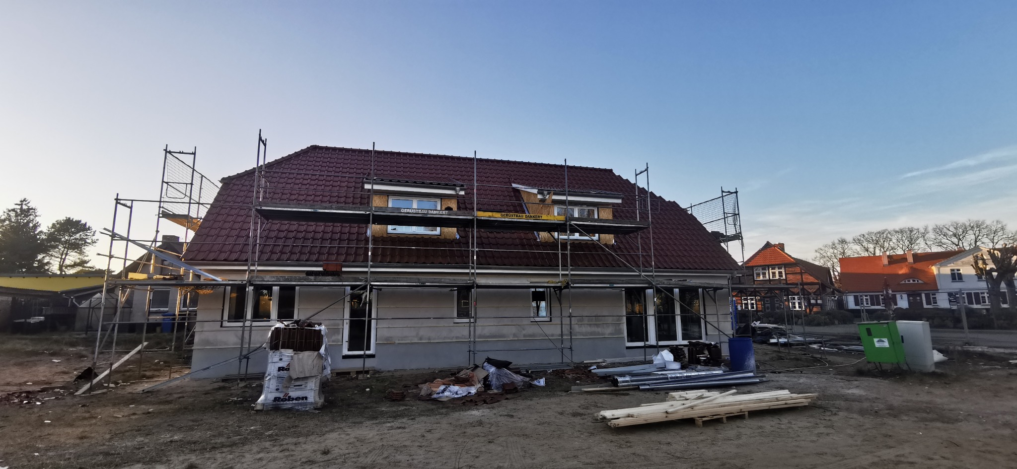 Dom w trakcie budowy z rusztowaniem, widoczne dachówki na dachu i materiały budowlane na ziemi. Ujęcie z zewnątrz, w tle inne budynki.