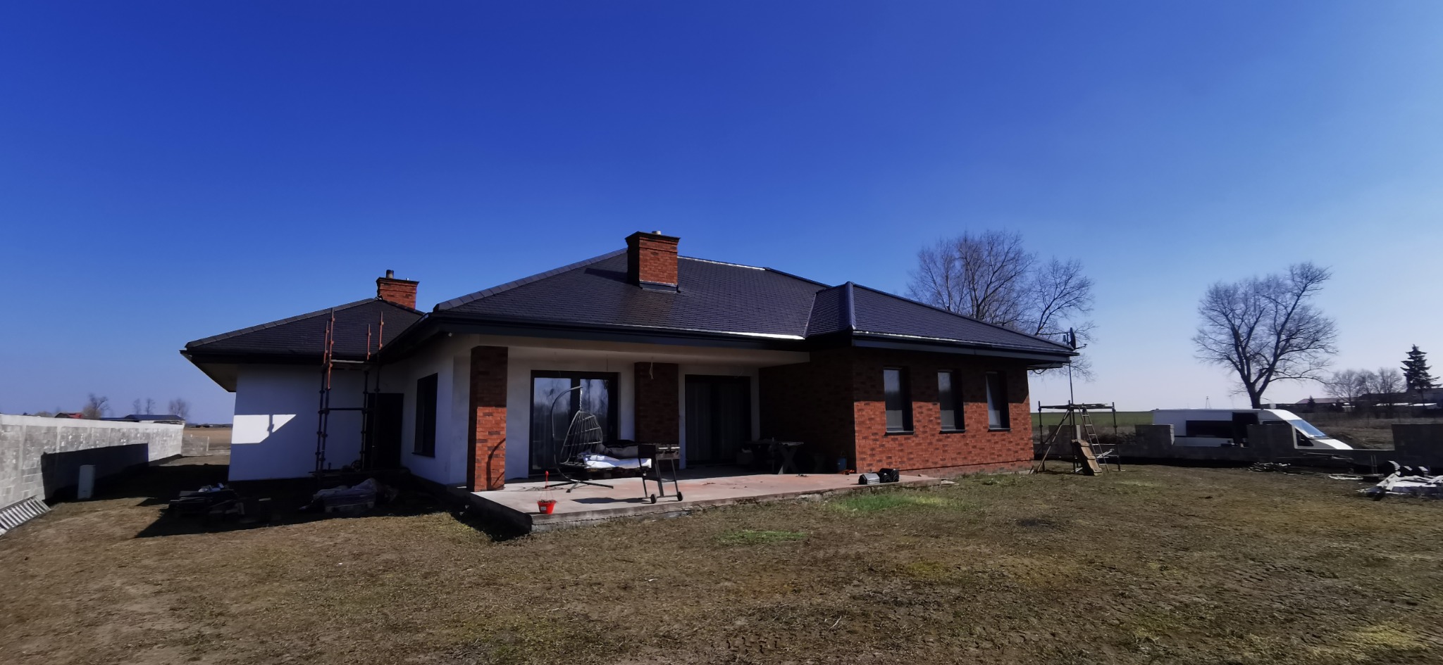 Nowoczesny dom jednorodzinny z ciemną dachówką i ceglanymi elementami elewacji, widziany z perspektywy ogrodu w słoneczny dzień. Widoczne rusztowanie przy ścianie.