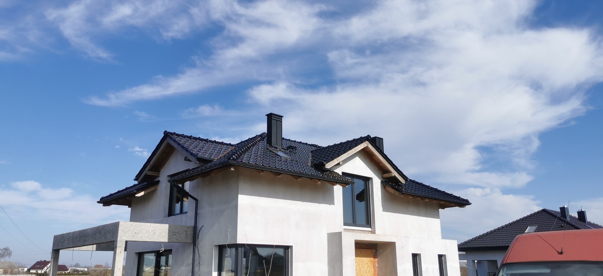 Dom jednorodzinny w trakcie budowy z nowym, błyszczącym dachem z czarnej dachówki ceramicznej i kominem na tle błękitnego nieba z chmurami.
