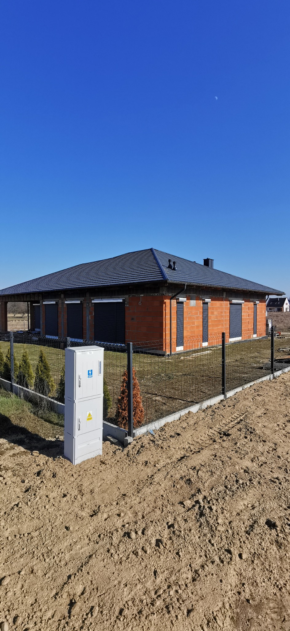 Niski, ceglany dom z ciemnoszarą dachówką ceramiczną, otoczony siatką ogrodzeniową i młodymi drzewkami iglastymi, na tle błękitnego nieba. Widoczna skrzynka elektryczna.